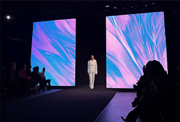 Model walking the WAVEY runway between two LED walls showing cyan and magenta fabric-like visuals